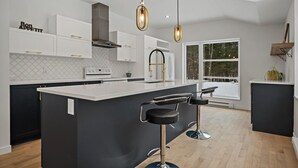 A modern kitchen with a central island, bar stools, pendant lighting, and a large window providing a view of the outdoors. - 1 month for the price of 1 week! (Mont-Tremblant)