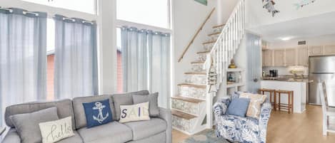 Una sala de estar con un sofá gris, un sillón estampado y una escalera que conduce a una cocina.