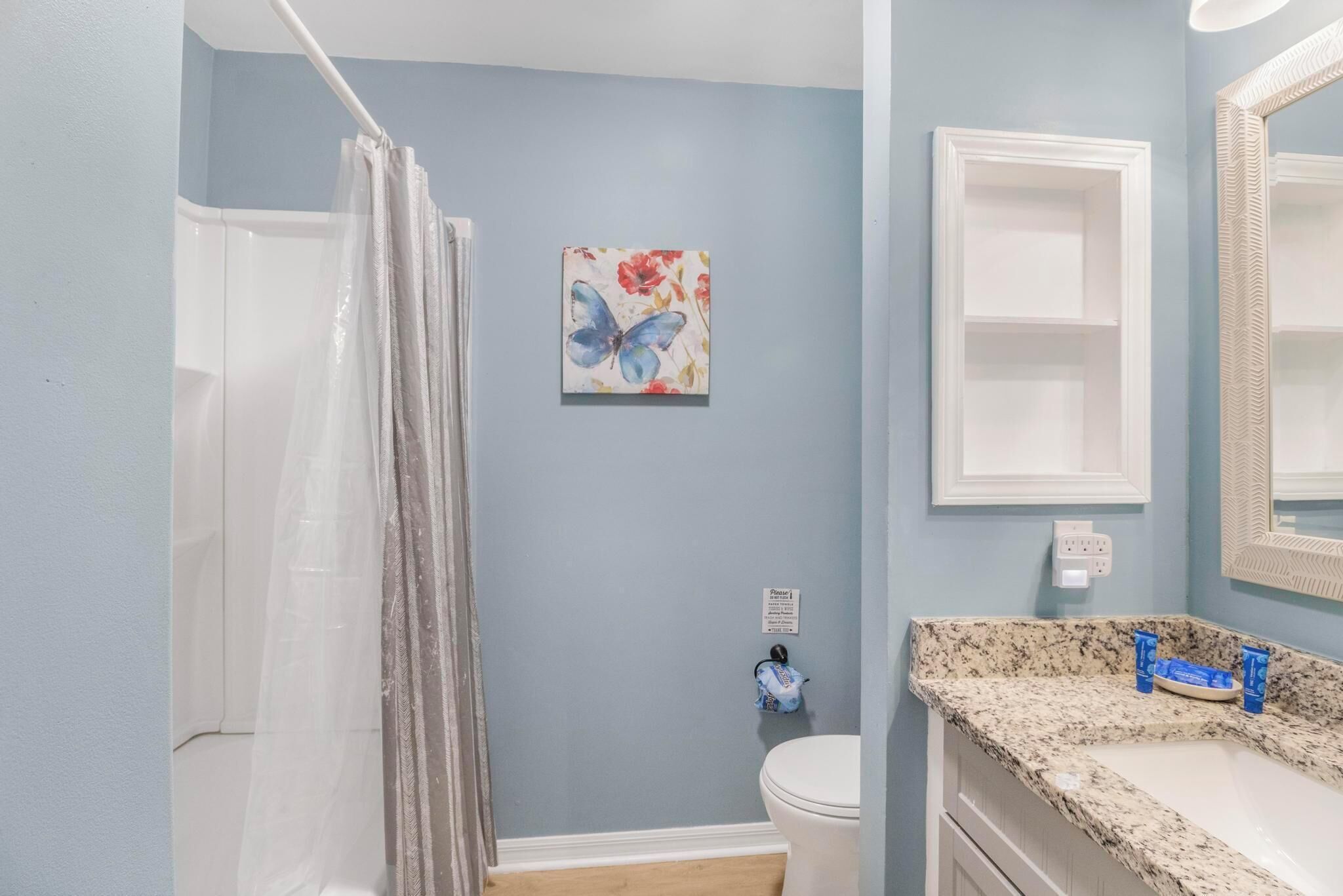 A bathroom with a shower curtain, a toilet, a sink with toiletries, and a wall-mounted mirror.