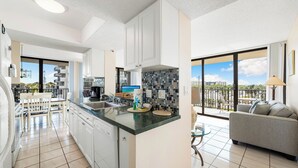 A modern kitchen with white cabinets, a granite countertop, and a large window offering a view of the outdoors. - Stunning Sunsets! 2 Bed, 2 Bath Condo on Siesta Key! (Siesta Key)
