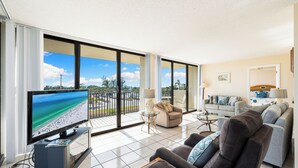 A modern living room with a large TV, comfortable seating, and a balcony with a view of the beach. - Stunning Sunsets! 2 Bed, 2 Bath Condo on Siesta Key! (Siesta Key)