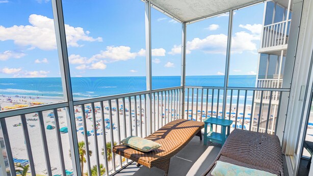 A balcony with a wicker sofa and a table, overlooking a beach with umbrellas and people. - Stunning Sunsets! 2 Bed, 2 Bath Condo on Siesta Key! (Siesta Key)