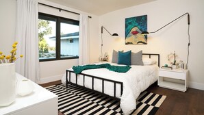 Un dormitorio moderno con cama, ventilador de techo, un cuadro y mesita de noche con lámpara.