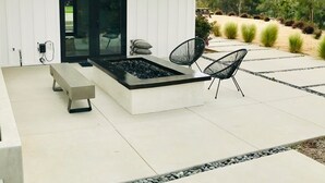 Un patio moderno al aire libre con un elemento decorativo de fuego rectangular, dos sillas negras y un banco.