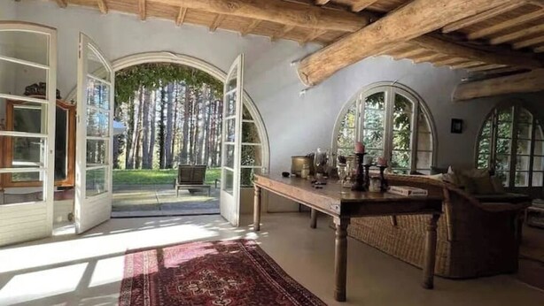 Una habitación amplia con un techo de madera grande, una mesa de comedor con sillas, un sofá y vistas a un bosque a través de puertas abiertas.