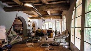 Un interior rústico con vigas de madera a la vista, una mesa de madera con diversos objetos y amplias ventanas arqueadas que dejan entrar luz natural.