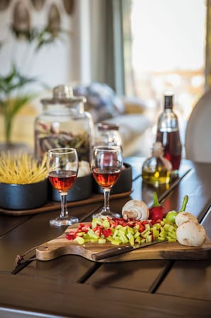 A wooden cutting board with chopped vegetables, two glasses of red wine, and a bottle of olive oil on a table.