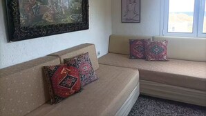 A living room with a beige sofa, patterned cushions, a framed landscape painting, and a window with a view. - Sunny apartment with view to the Mirabello Gulf (Agios Nikolaos)