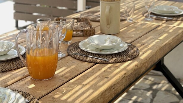 Ein Holztisch, draußen gedeckt für ein Essen, mit einem Glasbehälter mit Orangensaft, Tellern und einer gewebten Tischmatte.