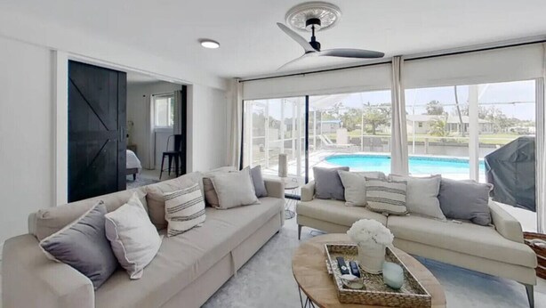 Ein geräumiges Wohnzimmer mit großem Fensterblick auf einen Swimmingpool, Deckenventilator und moderner Inneneinrichtung.