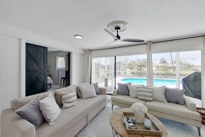 Un salon spacieux doté d’une grande fenêtre donnant sur une piscine, d’un ventilateur de plafond et d’un aménagement intérieur moderne.
