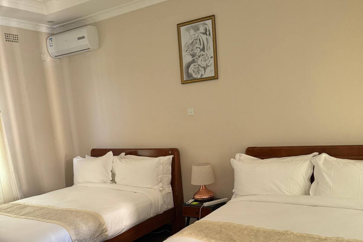 Quarto de hotel com duas camas, uma escrivaninha, uma cadeira, um abajur e um aparelho de ar condicionado.