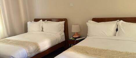 A hotel room with two beds, a desk, a chair, a lamp, and an air conditioning unit.