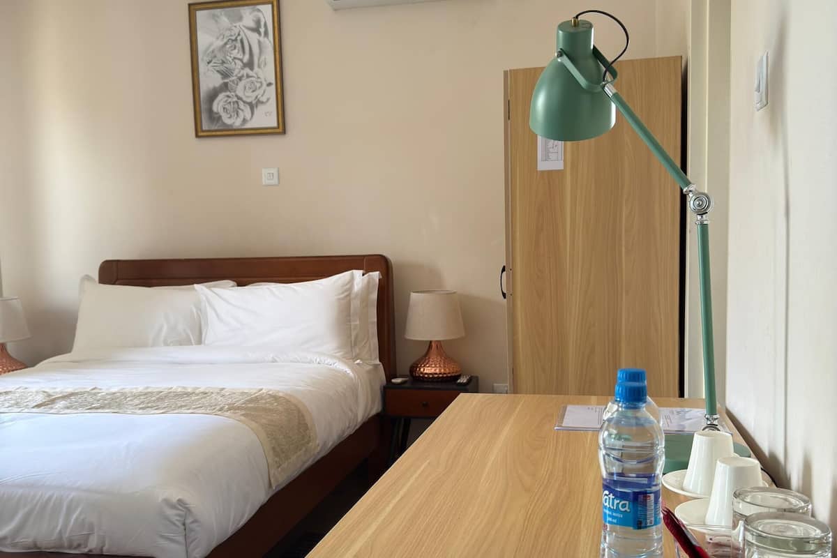 Quarto de hotel com cama, mesa de cabeceira de madeira, escrivaninha com garrafa de água, luminária e quadro na parede.