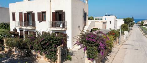 A residential street with white buildings, balconies, and purple flowering plants.