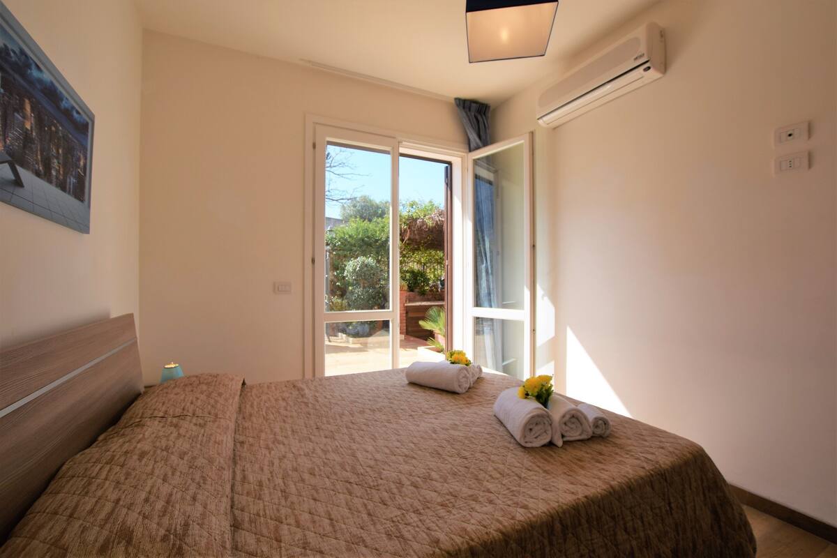 A bedroom with a large bed, a nightstand, an air conditioning unit, and a view of the outdoors through a sliding glass door.
