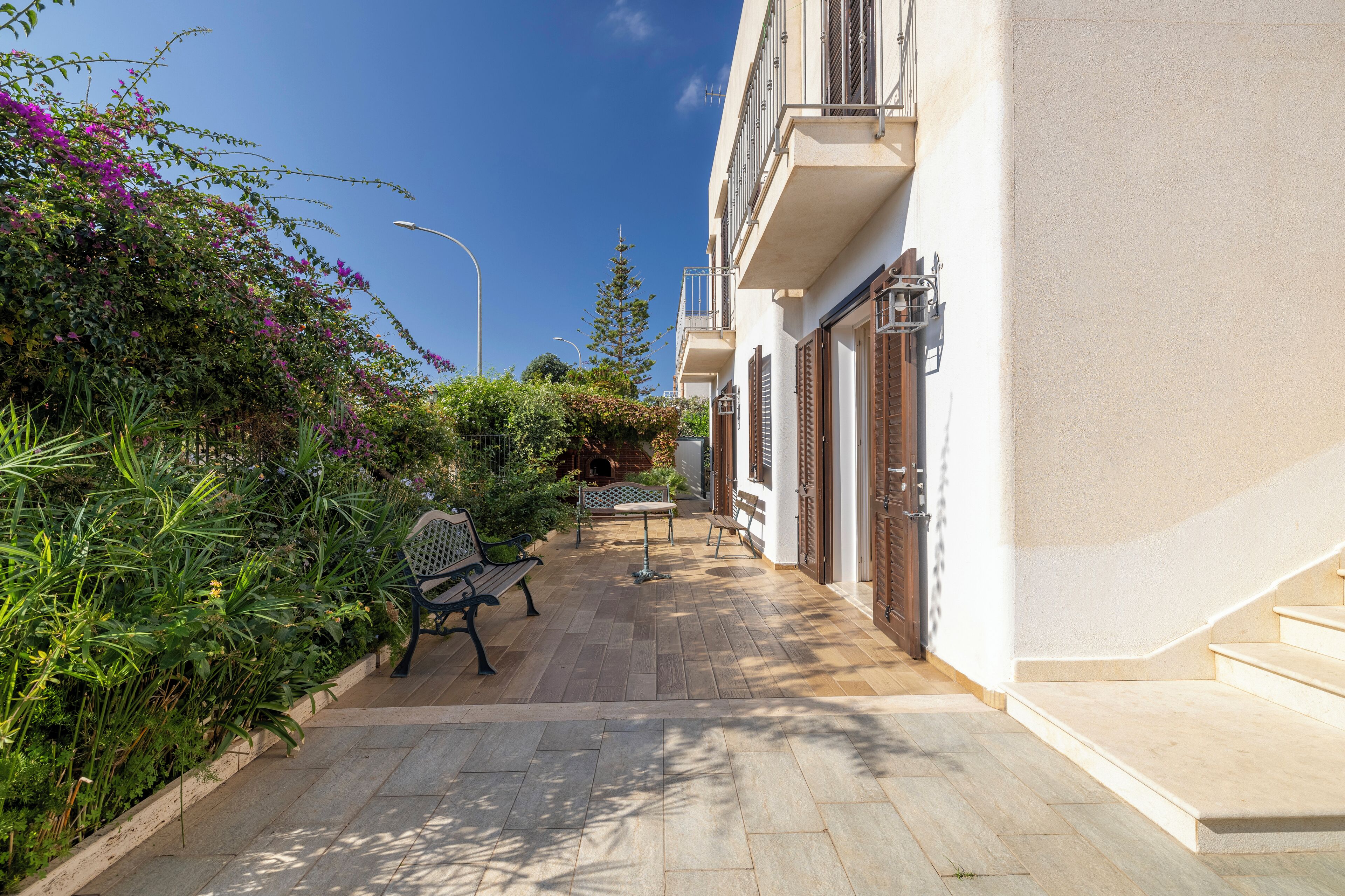A narrow outdoor corridor with a bench and a table, surrounded by lush greenery and a white building with balconies.