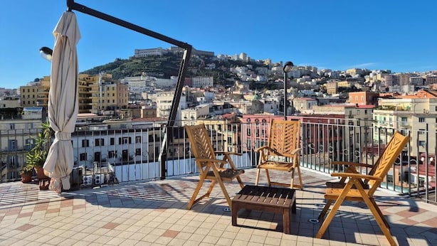 A rooftop terrace with wooden chairs and a table, overlooking a cityscape with buildings and a hill. - Appartamento Annunziata (Naples)