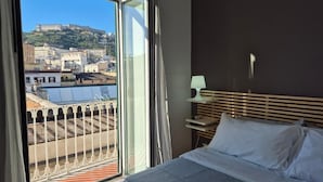 A hotel room with a bed, a nightstand, a lamp, and a view of a cityscape through a large window. - Appartamento Annunziata (Naples)