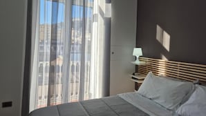 A hotel room with a bed, a nightstand with a lamp, and a view through a window with sheer curtains. - Appartamento Annunziata (Naples)