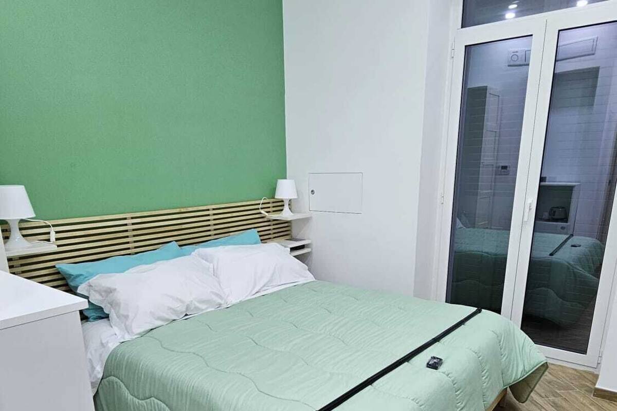 A bedroom with a bed, a nightstand, a lamp, a wardrobe, and a sliding glass door leading to a balcony.