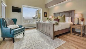 A bedroom with a large bed, a television, a chair, and a window with a view of a building.