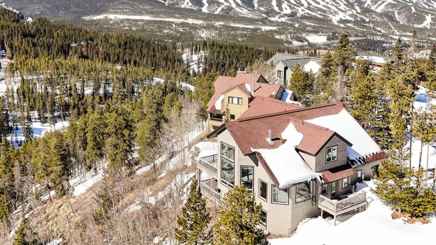 A large, modern house with multiple levels and a prominent balcony, surrounded by snow-covered mountains and dense forest.