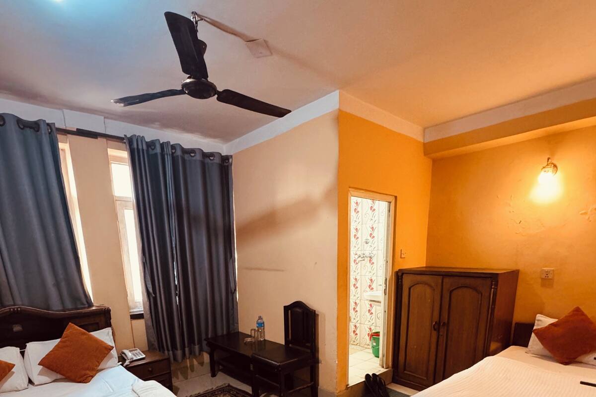 A hotel room with two beds, a ceiling fan, a desk with a chair, a wardrobe, and a window with curtains.
