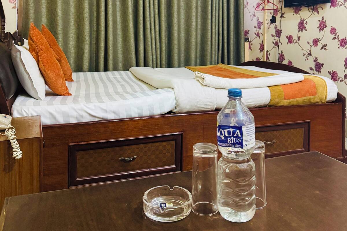 A hotel room with a bed, a table with a bottle and glasses, a telephone, and an air conditioning unit.