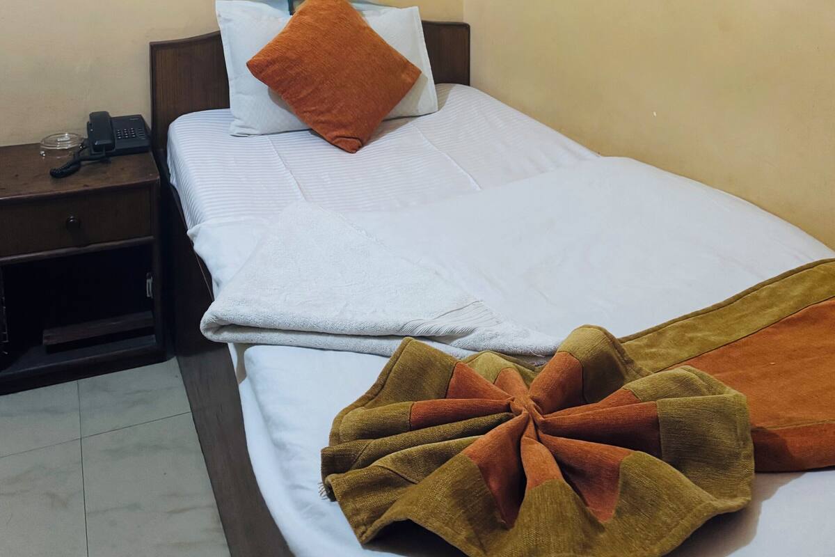 A single bed with a white and brown blanket, a brown pillow, and a bedside table with a phone and a glass.
