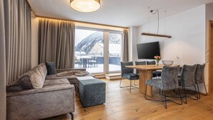 Ein modernes Wohnzimmer mit Sofa, Hocker und Essbereich mit Stühlen. Ein Fernseher ist an der Wand montiert und es gibt ein großes Fenster mit Bergblick.