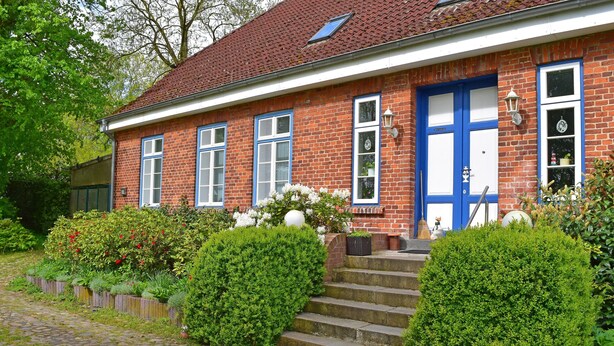 Ein rotes Backhaus mit blauer Tür und Fensterläden in Weiß, umgeben von gepflegten Hecken und einem Kopfsteinpflasterweg.