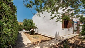 Un pequeño edificio blanco con una puerta y ventana, rodeado de follaje y un camino pavimentado.