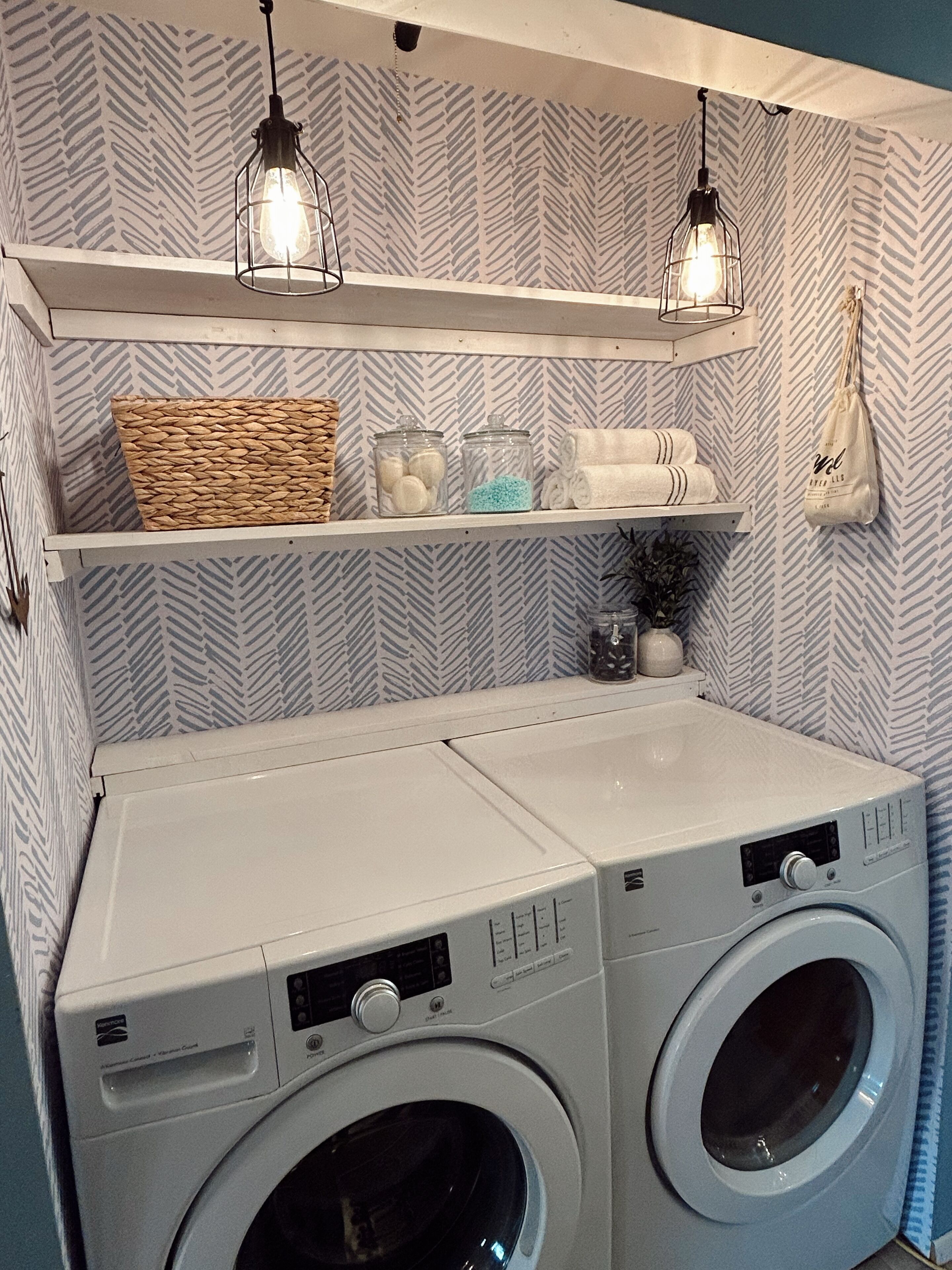 A laundry area with two front-loading washing machines, a white sink, and shelves above holding towels, a woven basket, and glass jars.