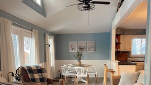 Un salon chaleureux avec un ventilateur de plafond, un coin repas avec une table et des chaises blanches, et une cuisine visible à travers une porte ouverte.