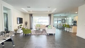 A modern living room with a sofa, armchairs, a coffee table, and a foosball table. There is a kitchen area with bar stools and a dining table. - Spacious 4-bedroom house in beautiful Cape Coral with WiFi, AC, Dock, Pool, Spa (Cape Coral)