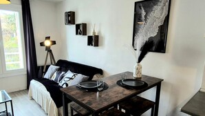 A modern living room with a dark wooden dining table, a black sofa with patterned cushions, a floor lamp, and a framed abstract painting. - L'Atelier-T2 near the Wifi-Parking center (Lourdes)