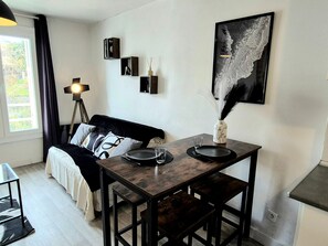 A modern living room with a dark wooden dining table, a black sofa with patterned cushions, a floor lamp, and a framed abstract painting. - L'Atelier-T2 near the Wifi-Parking center (Lourdes)