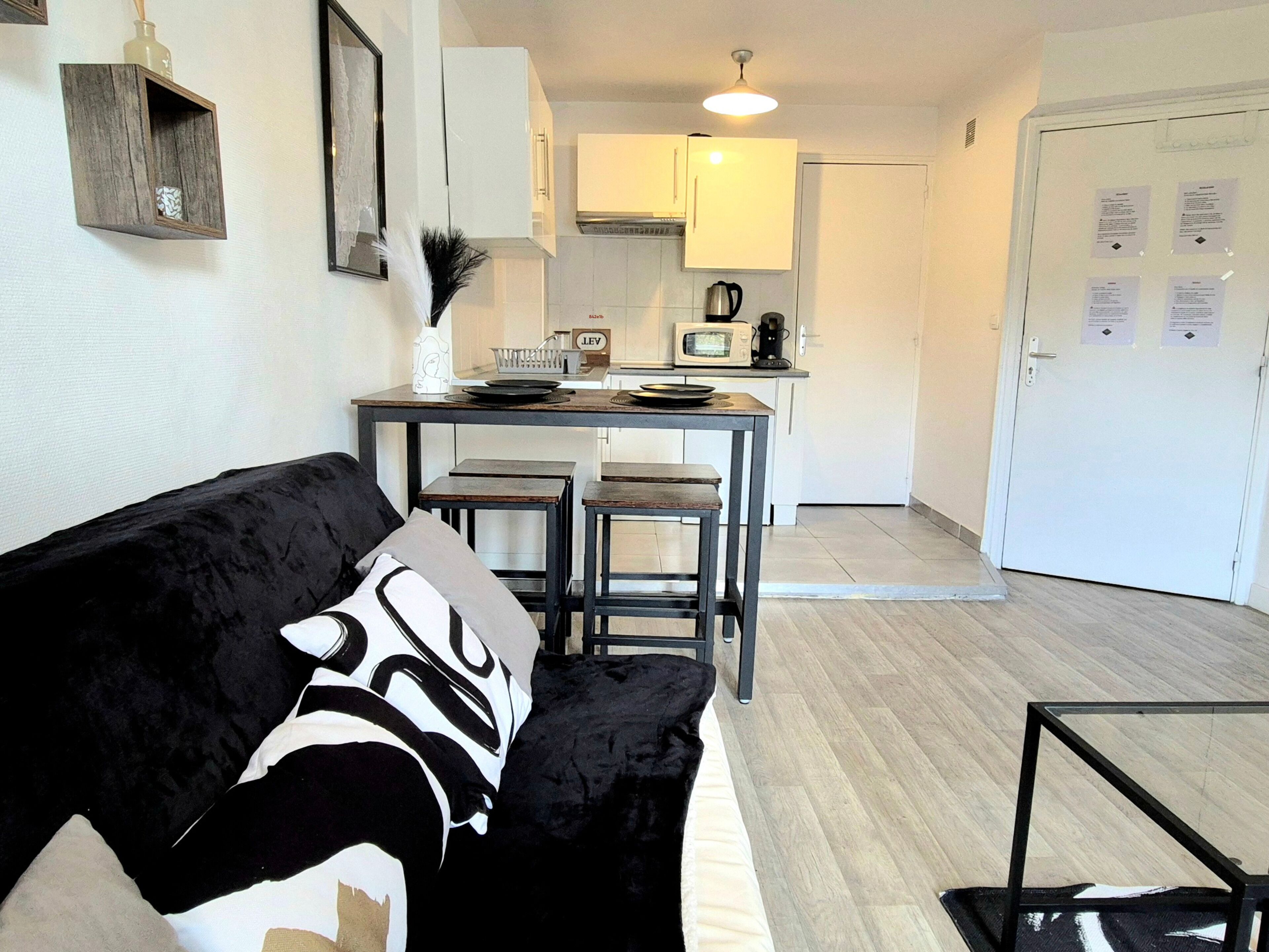 A living room with a black and white sofa, a small table, and a kitchen area with a microwave and cabinets.