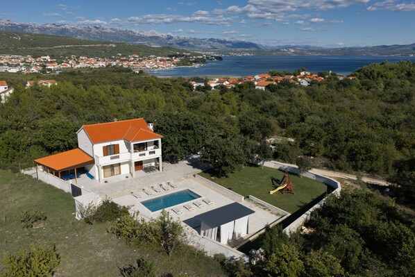 A modern two-story house with a swimming pool, surrounded by greenery and a view of the sea.