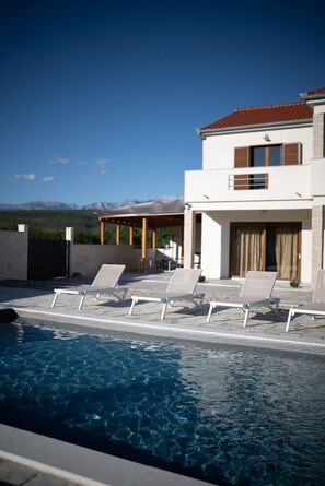 A modern two-story building with a swimming pool, lounge chairs, and a view of mountains.