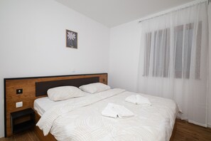 Une chambre à coucher avec une tête de lit en bois, un lit simple avec une literie blanche, une petite table de chevet, un interrupteur fixé au mur et une fenêtre avec des rideaux légers.