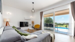 Un salon moderne avec un canapé gris, un téléviseur à écran plat, un fauteuil jaune et une grande porte-fenêtre coulissante donnant sur une piscine.