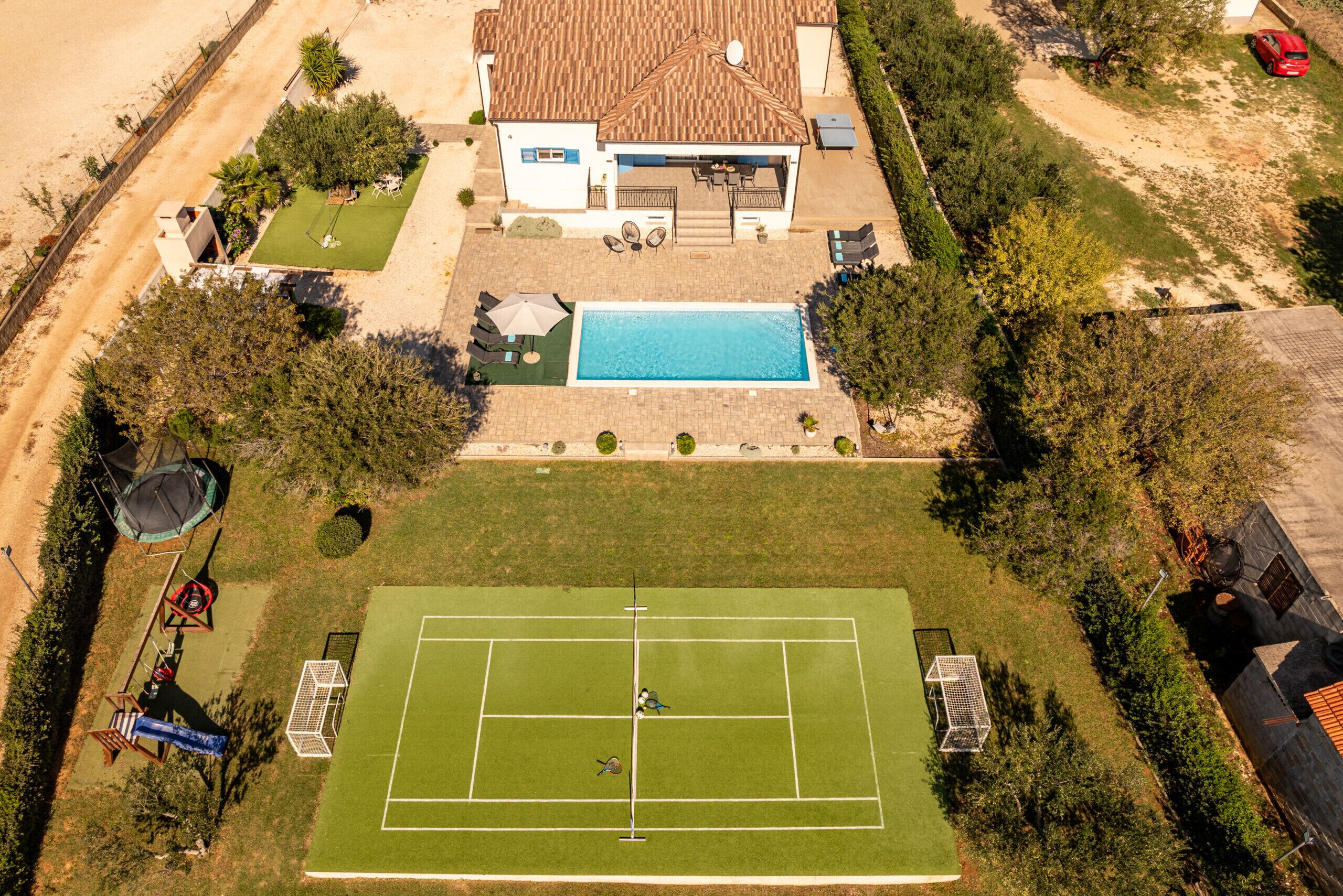 An aerial view of a residential property with a swimming pool, tennis court, and a fenced yard.