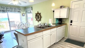 Une cuisine dotée d’un plan de travail en bois, de meubles blancs, d’une décoration en forme de roue de navire et offrant une vue sur l’océan grâce à une porte-fenêtre coulissante.