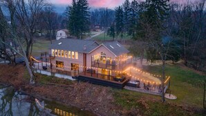 A two-story house with a deck and outdoor lighting, surrounded by trees and a body of water. - Riverside Retreat  Sleeps 20 (Halifax)