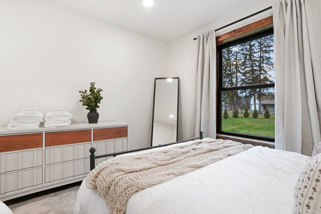 Une chambre à coucher comprenant un lit, une commode, un miroir et une fenêtre donnant sur des arbres et une pelouse.
