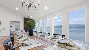 A dining area with a long table set for multiple guests, large windows offering a view of the sea, and a chandelier hanging from the ceiling. - Riverside Retreat  Sleeps 20 (Halifax)