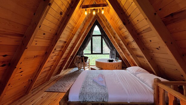 Ein gemütliches Schlafzimmer unter dem Dach mit Holzbalkendecke, einem großen Fenster, einer integrierten Badewanne und einem Bettgestell aus Holz.