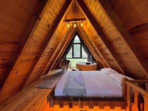 Une chambre confortable située sous les toits, avec un plafond en pente en bois, une grande fenêtre, une baignoire intégrée et un cadre de lit en bois.